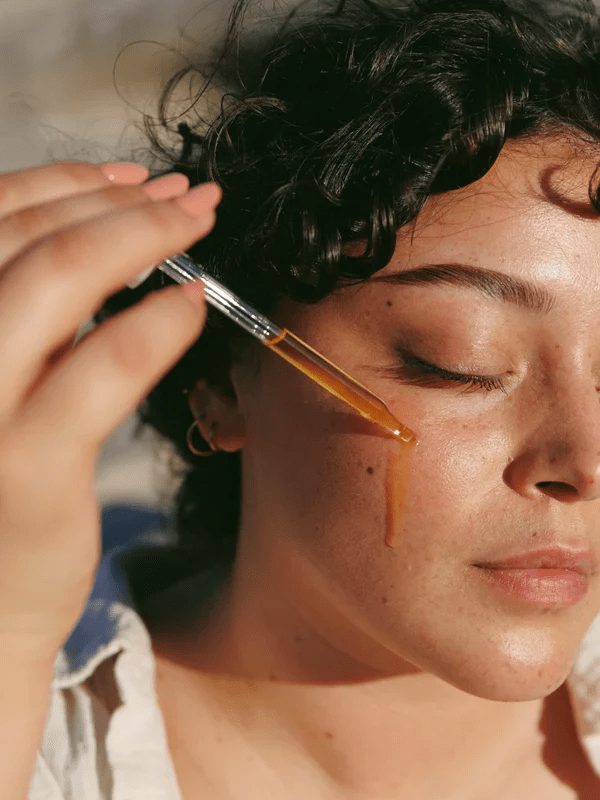 A woman dribbles a face oil on her cheek. Learn more about the best oil for your face by skin type in our complete guide.