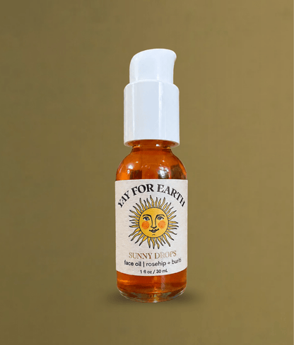 A small amber glass bottle of 'Sunny Drops' face oil by 'Yay For Earth' with a white pump dispenser, set against a solid olive-brown background.