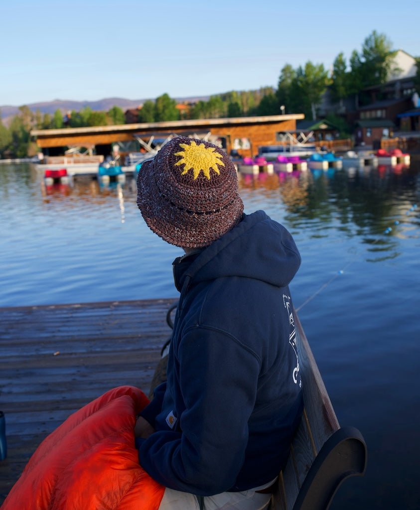 Person sitting on a dock by a lake wearing a hat with a sun design.