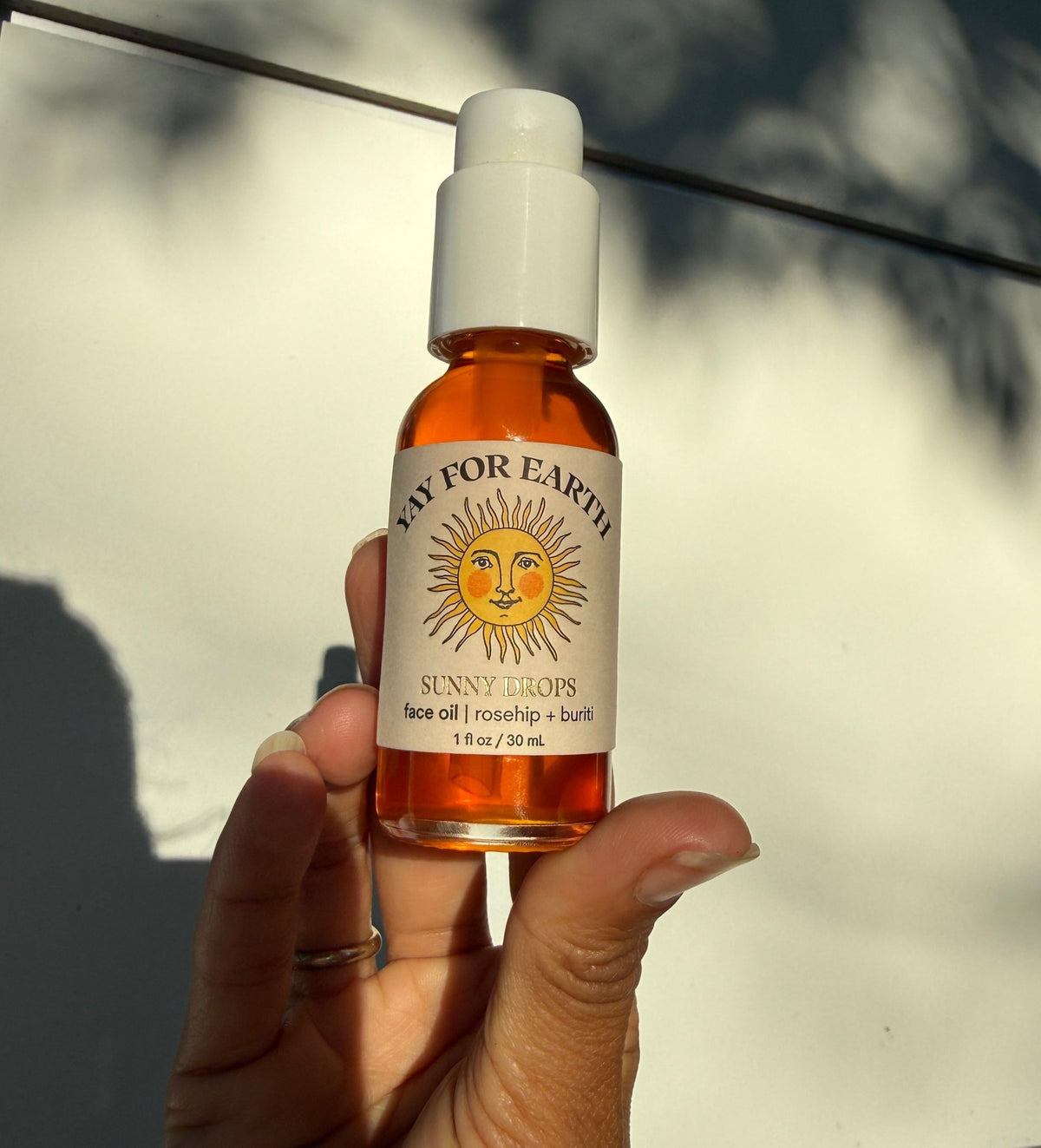 A hand holds a small clear glass bottle of face oil with a white cap and a cream label featuring a sun illustration. The label reads 'Yay For Earth' and 'Sunny Drops face oil'.