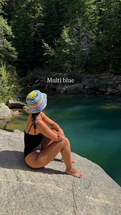 Person sitting on a rock by a pool of water with a colorful hat and black swimsuit.